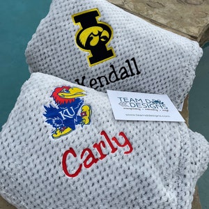 May include: Two white fleece blankets with embroidered logos and names. The top blanket has a black and yellow University of Iowa logo and the name "Kendall". The bottom blanket has a blue and red University of Kansas logo and the name "Carly".