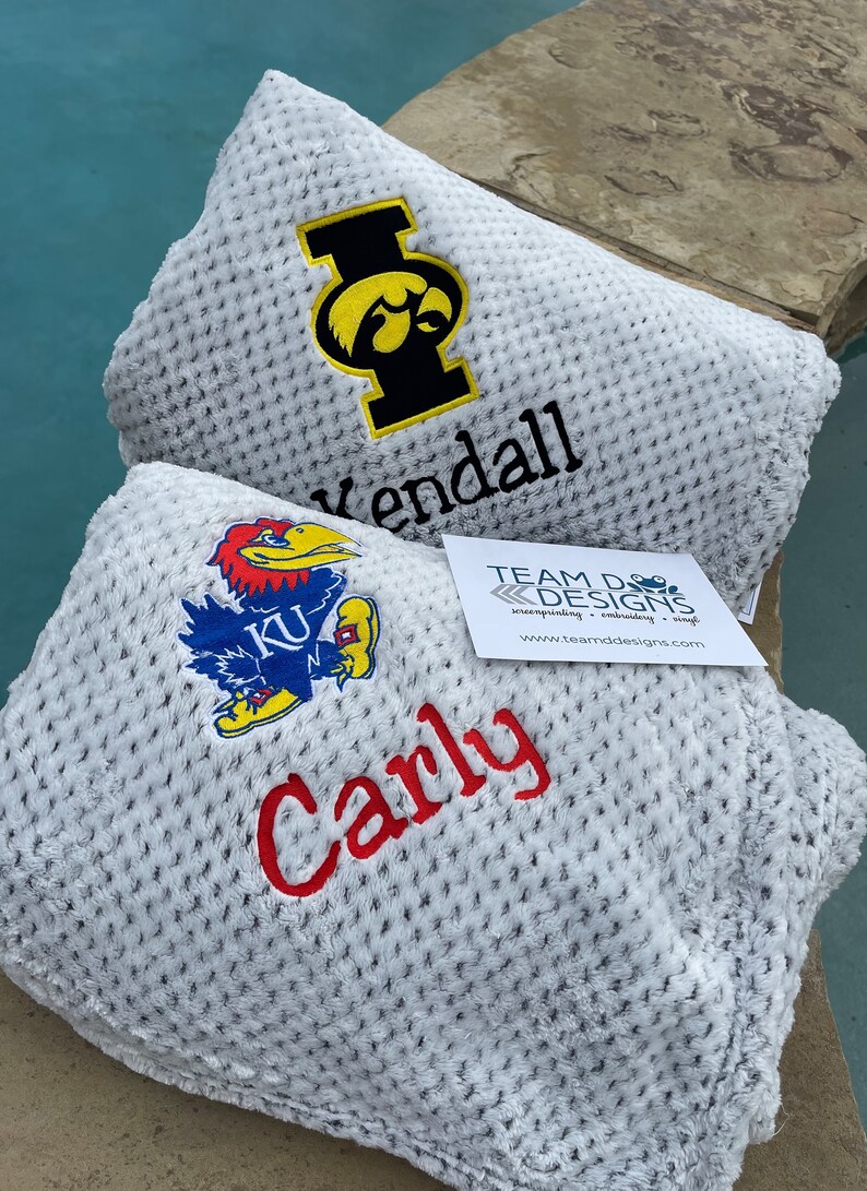 May include: Two white fleece blankets with embroidered logos and names. The top blanket has a black and yellow University of Iowa logo and the name "Kendall". The bottom blanket has a blue and red University of Kansas logo and the name "Carly".