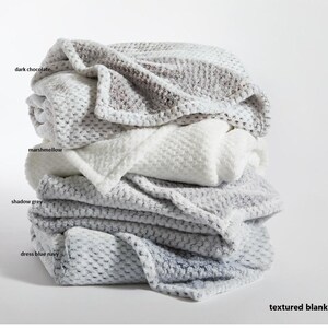 May include: A stack of four textured blankets in shades of gray, white, and blue. The blankets are labeled with the colors "dark chocolate", "marshmallow", "shadow grey", and "dress blue navy". The text "textured blankets" is at the bottom of the image.