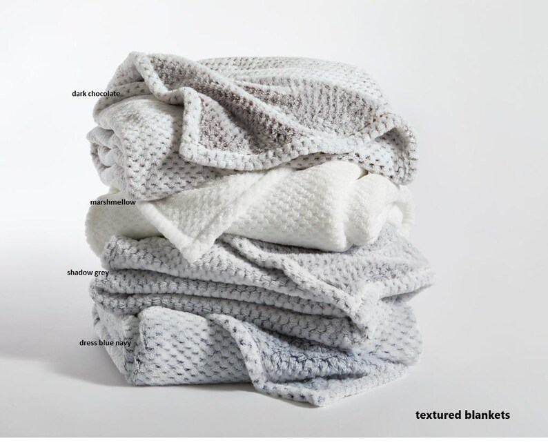 May include: A stack of four textured blankets in shades of gray, white, and blue. The blankets are labeled with the colors "dark chocolate", "marshmallow", "shadow grey", and "dress blue navy". The text "textured blankets" is at the bottom of the image.