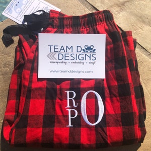 May include: Red and black buffalo plaid pajama pants featuring a white embroidered monogram with the letters R, P, and O. A white card with the text TEAM D DESIGNS, screenprinting, embroidery, vinyl, and www.teamddesigns.com is placed on top.