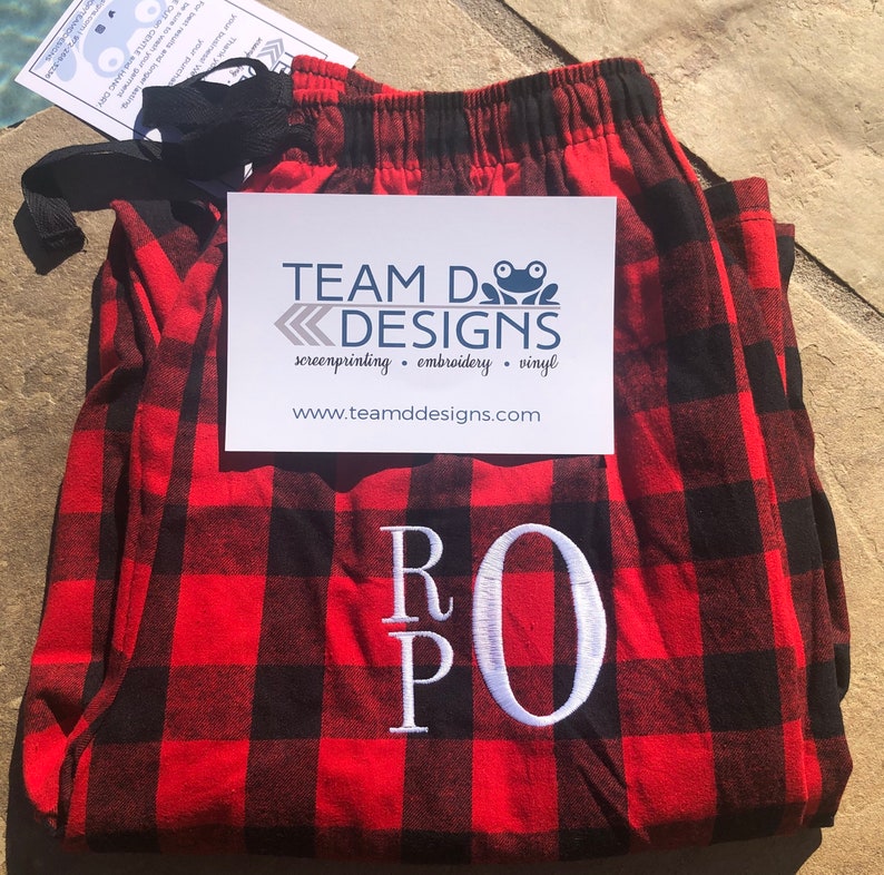 May include: Red and black buffalo plaid pajama pants featuring a white embroidered monogram with the letters R, P, and O. A white card with the text TEAM D DESIGNS, screenprinting, embroidery, vinyl, and www.teamddesigns.com is placed on top.