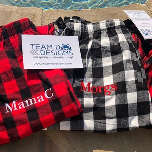 May include: Three pairs of flannel pajama pants featuring a buffalo plaid pattern. Two pairs are red and black, and one pair is white and black. Each pair is embroidered with a name: MamaC, Morgs, and Pook. A Team D Designs card is visible.