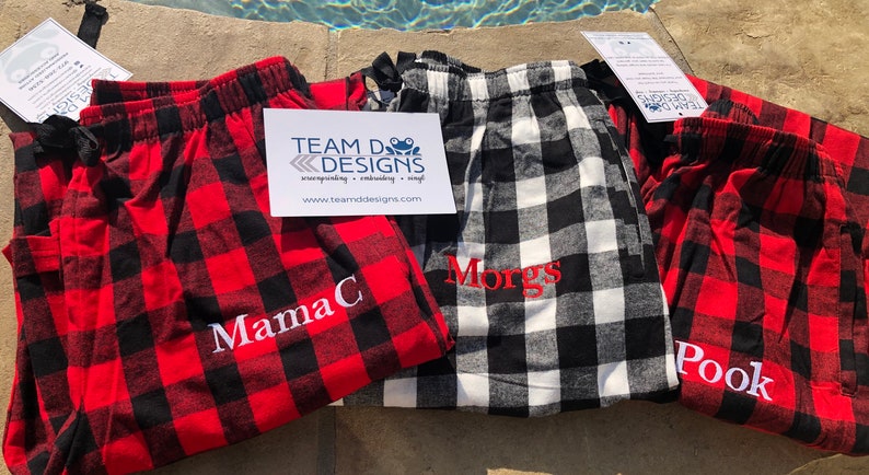 May include: Three pairs of flannel pajama pants featuring a buffalo plaid pattern. Two pairs are red and black, and one pair is white and black. Each pair is embroidered with a name: MamaC, Morgs, and Pook. A Team D Designs card is visible.