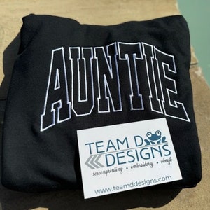 May include: Black sweatshirt with white embroidered text that reads "AUNTIE" in a large, bold font. A white business card with the text "TEAM D DESIGNS screenprinting. embroidery. vinyl www.teamddesigns.com" is visible in the bottom right corner of the image.