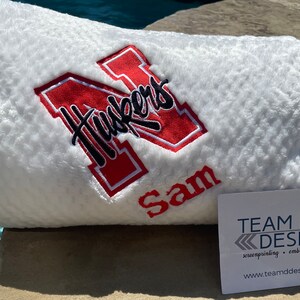以下が含まれることがあります： 赤い刺繍の「N」と黒い筆記体の「Huskers」が施された白いぬいぐるみブランケット。 「N」の下には「Sam」という名前が刺繍されています。