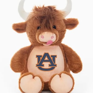 May include: A plush brown Highland cow stuffed animal with white horns and a pink tongue sticking out. The cow has a large "A" logo on its chest in navy blue and orange. The stuffed animal is sitting upright.