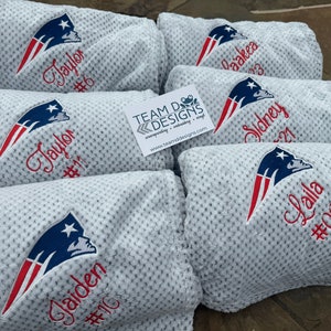 May include: Five white fleece blankets with the New England Patriots logo embroidered on them. Each blanket has a name and number embroidered below the logo. The names are Jaylor, Gaiden, Daakea, Sidney, and Lalla. The numbers are 6, 10, 3, 21, and 00.