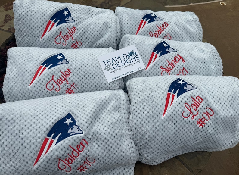 May include: Five white fleece blankets with the New England Patriots logo embroidered on them. Each blanket has a name and number embroidered below the logo. The names are Jaylor, Gaiden, Daakea, Sidney, and Lalla. The numbers are 6, 10, 3, 21, and 00.