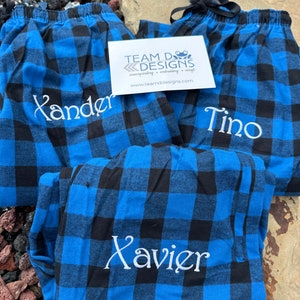 May include: Three pairs of blue and black buffalo plaid pajama pants featuring white embroidered names Xander, Tino, and Xavier. A white rectangular business card with the text TEAM D DESIGNS screenprinting embroidery vinyl is placed on top of the pants.