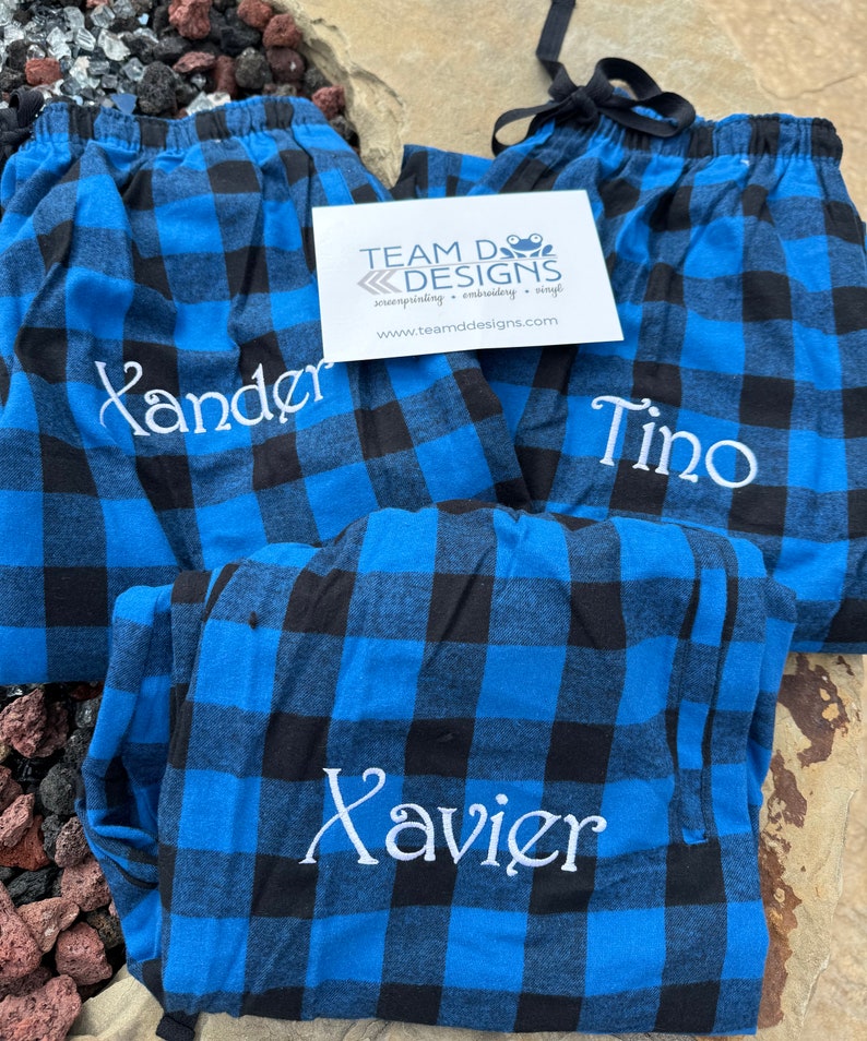 May include: Three pairs of blue and black buffalo plaid pajama pants featuring white embroidered names Xander, Tino, and Xavier. A white rectangular business card with the text TEAM D DESIGNS screenprinting embroidery vinyl is placed on top of the pants.
