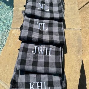 May include: A stack of four pairs of gray and black buffalo plaid pajama pants. Each pair features white embroidered monogram letters on the front, specifically reading NL, TL, JWH, and KHL from the top pair down to the bottom pair.