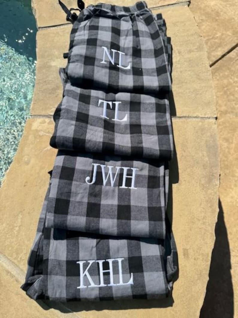 May include: A stack of four pairs of gray and black buffalo plaid pajama pants. Each pair features white embroidered monogram letters on the front, specifically reading NL, TL, JWH, and KHL from the top pair down to the bottom pair.