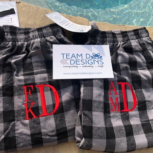 May include: Two pairs of gray and black buffalo plaid pajama pants with elastic waistbands and black drawstrings. Each pair features red embroidered monogram initials, FKD and AMD. A white card with the text TEAM D DESIGNS is placed on top.
