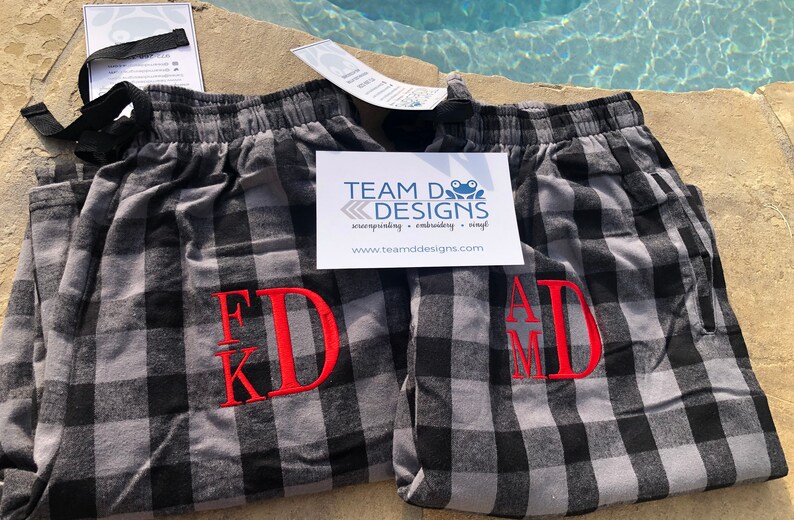 May include: Two pairs of gray and black buffalo plaid pajama pants with elastic waistbands and black drawstrings. Each pair features red embroidered monogram initials, FKD and AMD. A white card with the text TEAM D DESIGNS is placed on top.
