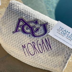 May include: A white, fuzzy blanket with a purple embroidered "ACU" and the name "MORGAN" below it. The blanket is folded and sitting on a brown surface. A business card for "TEAM DESIGN" is visible in the corner of the image.