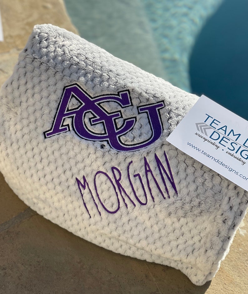May include: A white, fuzzy blanket with a purple embroidered "ACU" and the name "MORGAN" below it. The blanket is folded and sitting on a brown surface. A business card for "TEAM DESIGN" is visible in the corner of the image.