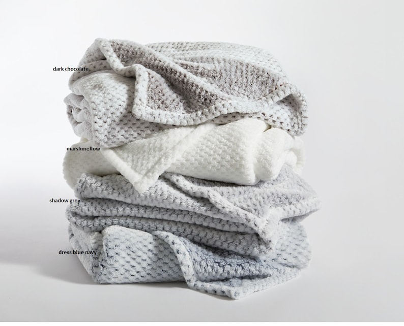 May include: A stack of four plush blankets in different colors: dress blue navy, shadow grey, marshmallow, and dark chocolate. The blankets are folded and stacked on top of each other.