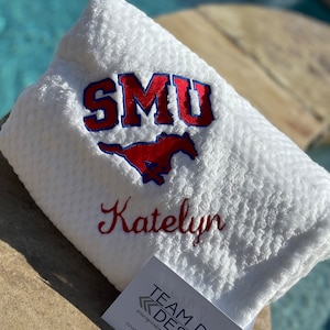 May include: White plush blanket with red and blue embroidered SMU logo and the name Katelyn. The blanket is folded and sitting on a brown surface.