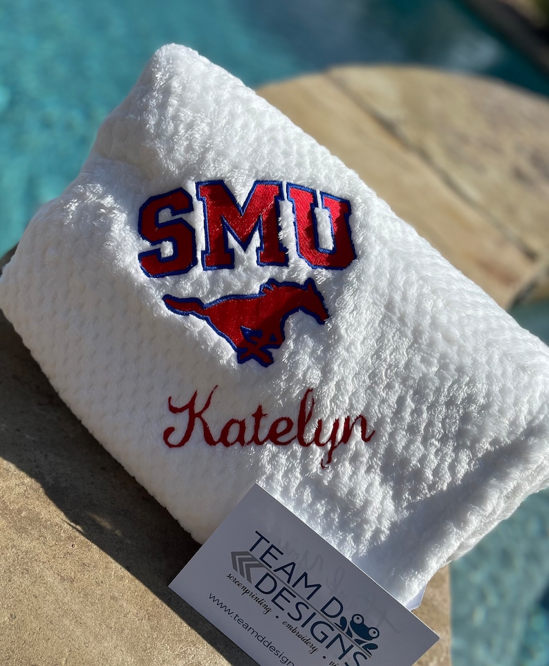 May include: White plush blanket with red and blue embroidered SMU logo and the name Katelyn. The blanket is folded and sitting on a brown surface.