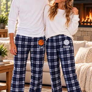 May include: Two pairs of matching navy blue and white plaid pajama pants. One pair features an orange basketball icon above the name Liat. The other pair features a white volleyball icon above the name Emma. Both are worn with long-sleeved white shirts.