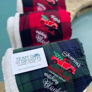 May include: Three fleece blankets with a red truck and Christmas tree design. The blankets are green, red, and blue plaid. The text on the blankets reads "Hallmark Christmas Movies watching blanket" and includes the names "Sharon's", "Kaje's", and a frog logo with the text "TEAM DDESIGNS screenprinting embroidery vinyl www.teamddesigns.com".