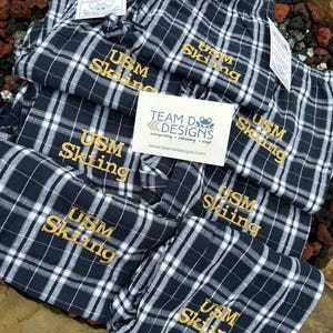 May include: A stack of black and white plaid pajama pants with the text "USM Skiing" embroidered in gold on each pair. The pants are folded and stacked on top of each other. A small white tag with the text "TEAM D DESIGNS" is visible in the lower left corner of the image.