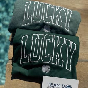 May include: Two green t-shirts with the word "LUCKY" embroidered in white on the front. Each shirt features a white shamrock design below the word.