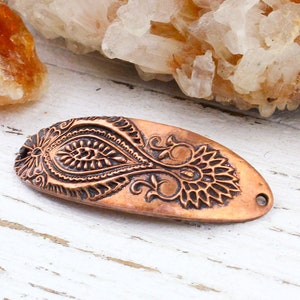 May include: Copper-coloured oval pendant with an embossed floral design. The pendant features a detailed paisley-like pattern with a small hole at each end. The background includes a white wooden surface and a piece of orange crystal.