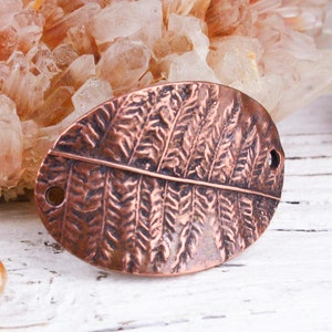 May include: Oval copper-colored metal pendant with a fern leaf design. The pendant has two holes and a textured surface. The background includes crystals and a pine cone on a white wooden surface.
