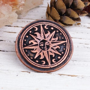 May include: A copper-coloured sun with a face and rays, surrounded by a black circle with a starburst design. The sun is on a white wooden surface.