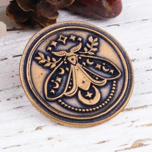 Peut inclure: Une épingle ronde de couleur bronze avec un motif détaillé d'un papillon de nuit aux ailes déployées. Le papillon de nuit est entouré d'étoiles, de lunes et de feuilles. L'épingle a un fond bleu foncé et des accents dorés.