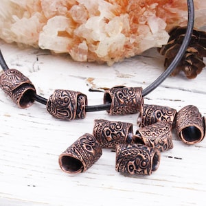 May include: A collection of cylindrical copper-toned beads featuring intricate embossed floral and scroll patterns. Several beads are threaded onto a thin black cord, while others are arranged in a cluster, showcasing the detailed metalwork.