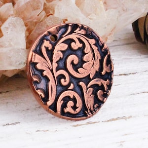 May include: A circular, copper-toned pendant with an elaborate, raised floral pattern. The pendant has a dark, oxidised background that highlights the detailed design. It features a small hole at the top for stringing. The pendant is approximately 3.8 cm in diameter.