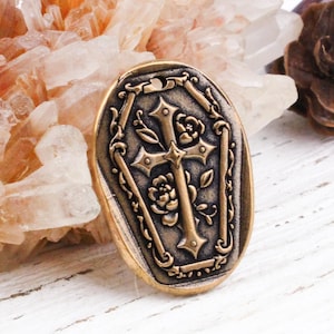 May include: An antique gold-colored wax seal with a cross and rose design. The oval seal has a decorative border and is set against a background of crystals and a white wooden surface.