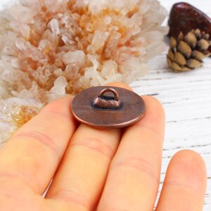 Pu&ograve; includere: Un bottone rotondo color rame con un anello sul retro. Il bottone &egrave; tenuto in mano, con uno sfondo sfocato di cristalli e una pigna. Il bottone &egrave; probabilmente per cucire o fare artigianato.