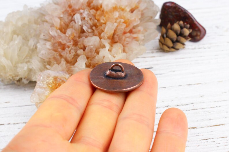 Pu&ograve; includere: Un bottone rotondo color rame con un anello sul retro. Il bottone &egrave; tenuto in mano, con uno sfondo sfocato di cristalli e una pigna. Il bottone &egrave; probabilmente per cucire o fare artigianato.