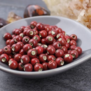 May include: A bowl filled with red ceramic beads. The beads have a speckled finish and a white glaze on the inside of the hole.