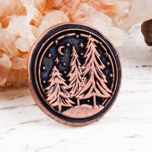 May include: A circular copper-toned metal pin featuring a raised relief design of three evergreen trees against a dark background. The scene includes a small crescent moon and several stars in the sky, all enclosed within a thin circular border.