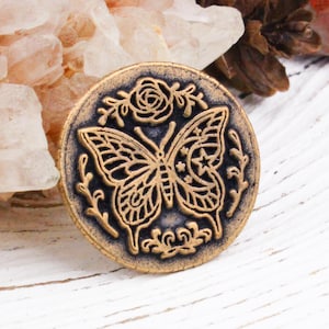 May include: A circular gold-toned metal button featuring a raised, dark-toned relief design of a butterfly centered between a rose at the top and leafy sprigs at the bottom. The butterfly wings are detailed with small star and crescent moon motifs.