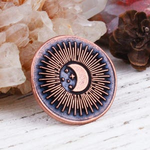 May include: A copper coin with a celestial design featuring a crescent moon and stars within a sunburst pattern. The coin has a blue-grey border and is displayed with crystals and a pinecone on a white wooden surface.