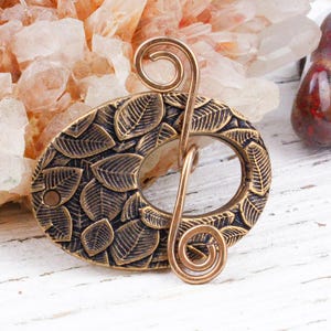 May include: An oval-shaped bronze hair barrette with a leaf design. The barrette has a decorative pin with a spiral design. The barrette is approximately 3 inches long and 2 inches wide.