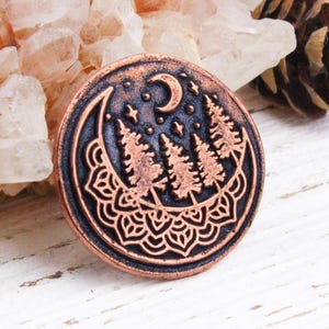 May include: A round, copper-colored metal pendant with a nature-inspired design. The pendant features a crescent moon, stars, and pine trees. The outer edge has a decorative mandala pattern. The pendant is displayed on a white wooden surface.