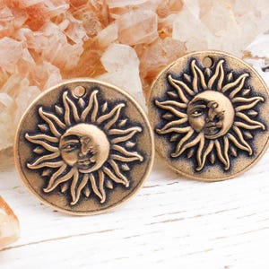 May include: Two circular bronze-toned metal charms featuring a raised relief design of a sun and crescent moon face. Each charm has a small hole at the top for attachment, with a dark antique finish highlighting the intricate facial features and rays.