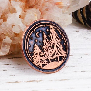 May include: A round, copper-colored metal pin with a detailed forest scene under a starry night sky. The design features silhouetted trees, a crescent moon, and stars, set against a dark blue background. The pin has a raised edge.