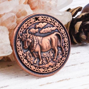 May include: A white ceramic mug with a black handle and the text "But First, Coffee" in a bold, black font. The mug is set against a blurred background. The mug appears to be approximately 10 cm tall.