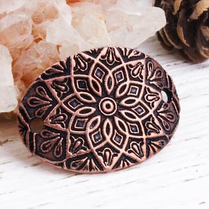 May include: An oval, copper-toned metal embellishment with an ornate floral design. The decorative piece features a central flower motif surrounded by geometric patterns. Two small holes are visible on either side of the oval.