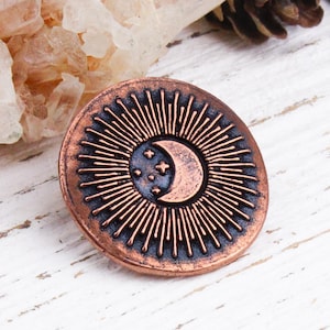 May include: A circular, copper-toned pin with a celestial design. It features a crescent moon and three stars at the centre, encircled by radiating lines. The design is set against a dark blue background.