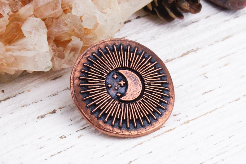 Pu&ograve; includere: Una spilla rotonda color rame con un design celeste. La spilla presenta una luna crescente e tre stelle al centro, circondate da linee radiali. Il design &egrave; posto su uno sfondo blu scuro.