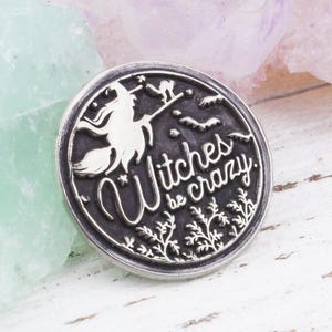 May include: A round, silver-colored pin with the phrase "Witches be crazy" in a stylized font. The pin features a witch on a broom, a cat, and decorative elements. The pin is set against a background of green and purple crystals.